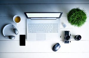 Top view of a table with a laptop, coffee mug, wireless headphones, cell phone, mouse, camera with an extra lens and a small round plant.