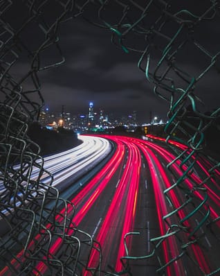 In the foreground there is a chain-link fence with a hold in it. Through the hole there are Traffic headlights on the highway at night with a city skyline in the background. In the foreground there is a chain-link fence with a hold in it