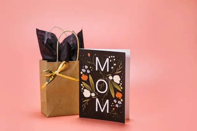 Pink background with a gold gift bag and a black card with flowers that says Mom