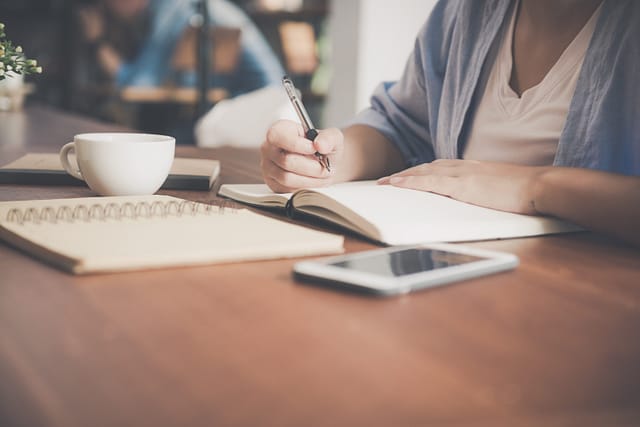 A person sitting a a wooden desk writing on a notebook. There is coffee and an iphone on the desk